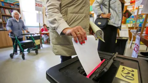 Getty Images A woman puts a piece of white paper into a black box at a polling station. She is wearing a cream coat and has painted her nails bright pink. The polling station seems to be in a children's library. There is a book case against the wall with lots of colourful books on display. 