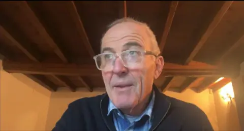 A man wearing glasses and wearing a blue short and top sits in room with a wooden beamed roof