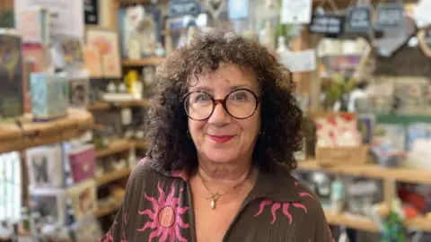 A woman standing in a shop looking at the camera and smiling. She is wearing glasses and a brown top with pink suns on it.
