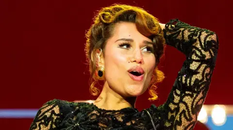 Raye smiles as she performs on stage in a black ballgown