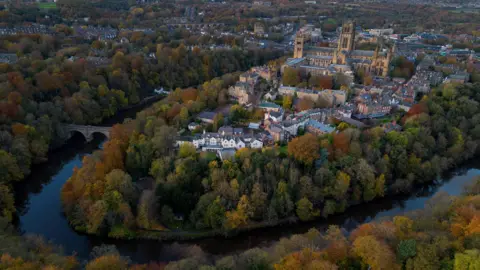 Reuters An aerial image of the city of Durham with the river Wear running around the area where the cathedral is. The river is lined by trees with autumnal colours. The cathedral is very prominent in the centre of the shot, towering over the surrounding buildings.