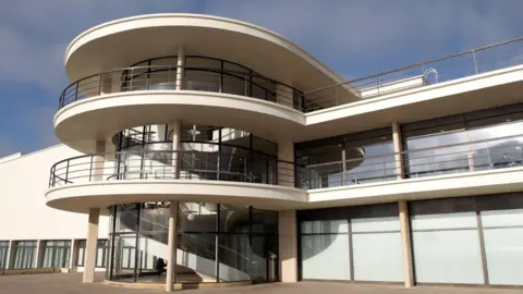 The De La Warr Pavilion’s modernist architecture has curved white balconies, with metal railings and a glass‑fronted stairwell with a spiral staircase inside. There are large rectangular windows looking out on to the open paved terrace in the foreground.