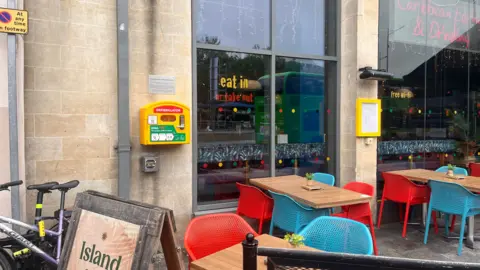 A yellow defibrillator is pictured on the exterior wall of a bar. There are tables and chairs, in red and blue, in front of two large plate-glass windows. 