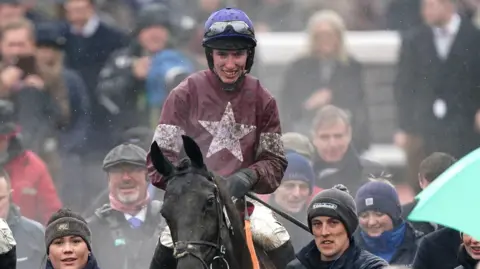 Jockey Jack Kennedy rides towards the camera on a black horse, with mud on his clothing and face