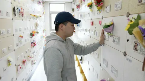 BBC Corrie/Junigin Choi is a man in a mask of gray masked jacket and a bispol hat stands in a memorial, surrounded by a flower of flowers, and holds his hand on a small image on the wall