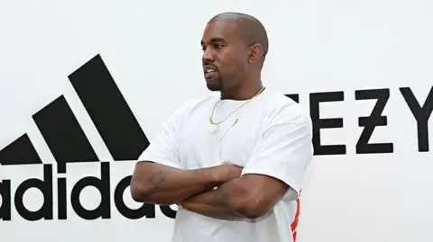 Getty Images Kanye West in a white T-shirt and gold chain poses for pictures with his arms crossed in front of the Yeezy and Adidas logo 