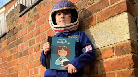 A child wearing blue overalls with an American flag stitched on the arm and a papier-mâché helmet. He is holding a book about American astronaut and physicist Sally Ride, and leaning against a brown brick wall outside, looking slightly down at the camera.