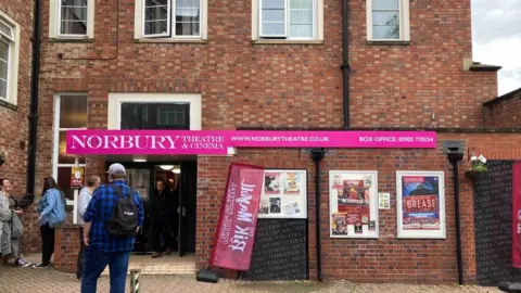 Two storeys are visible in this photo taken from the outside of the theatre. A pink and white sign above an entrance says Norbury Theatre & Cinema. Five people are standing outside.