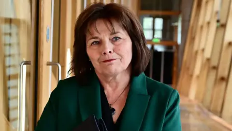 Getty Images Jeane Freeman in green dress jacket, walks along a corridor in the Scottish Parliament. She has medium-length dark hair.