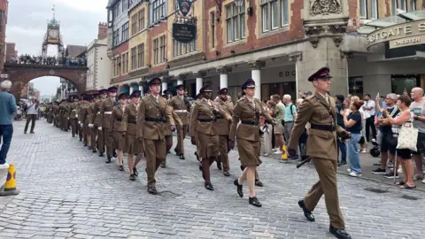 Soilders dressed in brown unifroms march on the high street, with crowds watching and taking pictures.