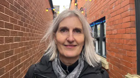 Adele looking into the camera, she's wearing a black jumper, a grey scarf, and a black winter coat. She's stood in one of Oswestry's side streets, you can see Christmas lights above her head and a shop window to her left.