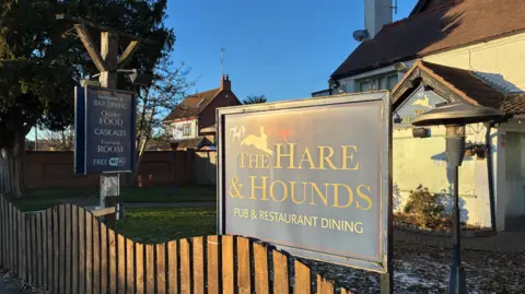 A pub site with a wooden fence in the foreground. There are two boards near the fence and a building is behind them.