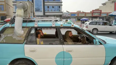 A light-blue Afghan taxi is with a hand-made air-cooler system fixed on top of it, - with its exhaust tube taped onto the window - passing through a market in Kandahar on 8 July. The driver is seen ferrying a male passenger. 