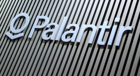 Close-up of the Palantir logo sign in white lettering mounted on a wall with vertical wooden slats in alternating light and dark tones.