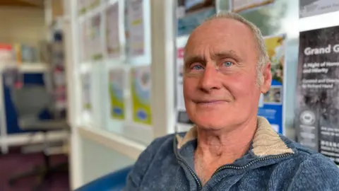 Michael Pye looking at the camera in a library with posters in the window behind him. He is wearing a blue jumper and smiling