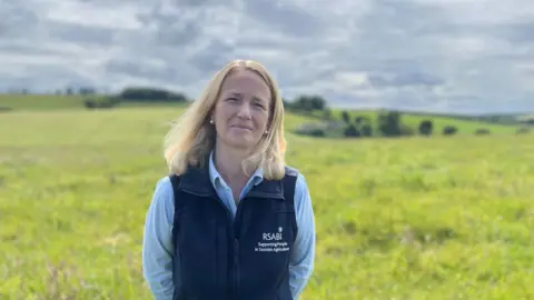A blonde-haired woman with a dark gilet and blue blouse looks at the camera with fields in the background
