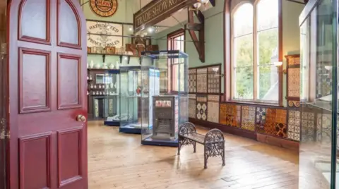 PA Media A wood-floored room with one large window and a number of glass display cases and multi-coloured tiles on the wall