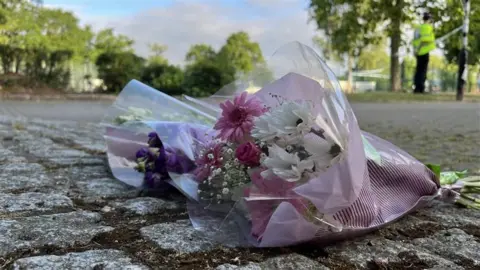 Stuart Woodward/BBC Bunches of white and purple flowers, still in their wrapping, are left on cobblestones in the park. In the distance you can see police tape around trees and one police officer standing by a post wearing a high vis jacket, black hat and trousers.