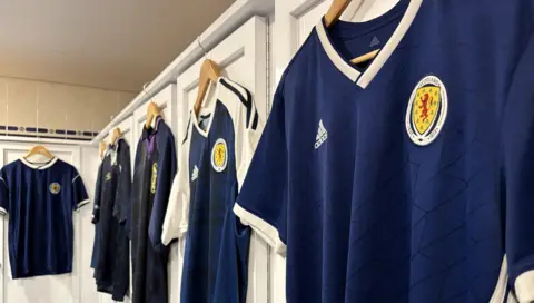 A row of Scotland jerseys over the years taken at the Scottish football museum. They are all hung up along the wall in a mock-up dressing room.