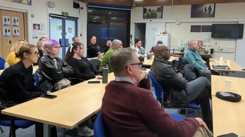 A group of people sit behind desks all facing towards one direction, as if watching a screen. 