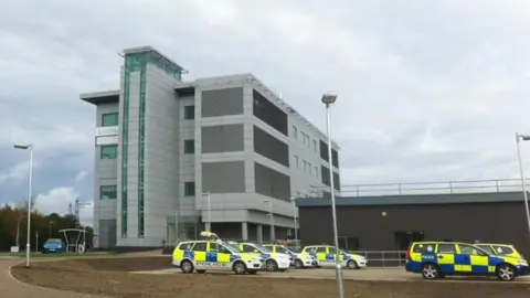 A view of Landmark House in Ipswich. The multi-storey building sits slightly to the left of the image on a grey overcast day. It is a grey building with several windows. Several police cars are parked in a car park at the front of the building. 