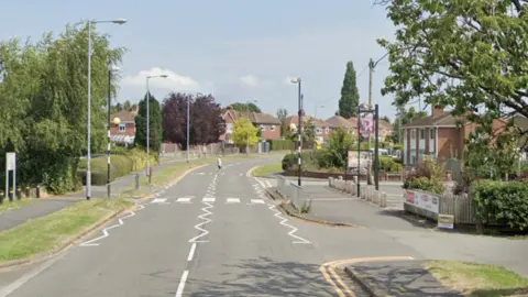 A suburban street lined by estate housing with trees either side and a pedestrian crossing visible