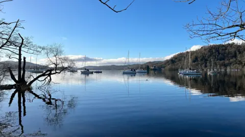 A general view of Ferry Nab at Windermere. The sky is blue with a few clouds reflecting on the water, which has just a few ripples. Lots of sailing boats are on the water and the ferry terminal is visible in the distance.