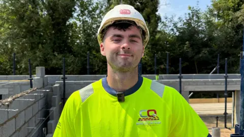 Cian McLoughlin with a white hard hat on, black hair, a yellow high-vis top on whilst on a building site.