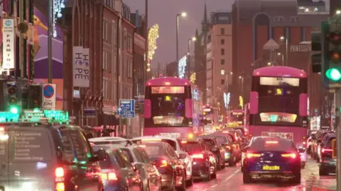 A long line of cars and buses are at a standstill in Belfast city centre. it is evening and all the brake lights on the vehicles are on.