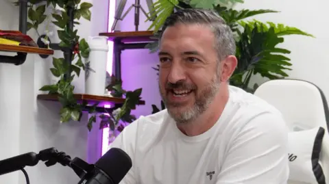 Andrew Williams A man with grey-black hair and a beard in a white long sleeved top, sitting behind a black microphone in a white-walled room