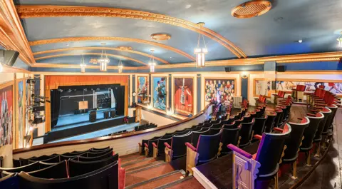 View from the balcony of the Electric Palace auditorium with a blue and gold vaulted ceiling, red velvet seats and orange curtains around the stage. The walls are decorated with murals of theatregoers.