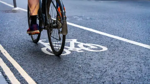 A cyclist riding in a designated bike lane marked by white lines and a painted bicycle symbol on a road. Only the bike’s rear wheel and the rider’s legs and shoes are visible.