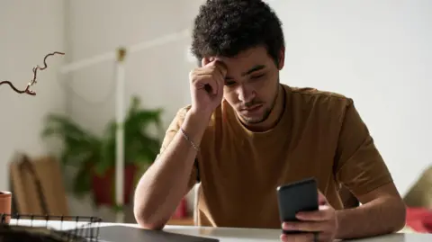 A young man looks at his phone while sitting at a computer in his home. He looks weary.