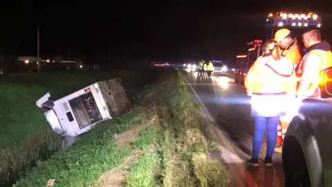 John Devine/BBC The bus on its side in a ditch. In the foreground are people in hi-vis jackets and a recovery van.