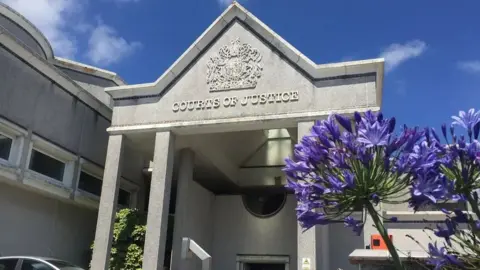 BBC The entrance of a grey court building, with an angular roof and concrete columns. A royal crest and the words Courts of Justice sit above the entrance and purple flowers are in front of the building.