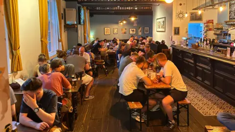 Mark Rackham Groups of men and women sit around tables in a pub. The bar is on the right, some are hunched over looking at the quiz answer sheet. 