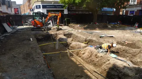 Colchester Archaeological Trust An archaeological dig taking place in Colchester's St Nicholas Square. There are several trenches dug into the ground, in which a man is on his hands and knees, wearing a high-visibility jacket and white safety hat. There are pieces of wood and tape running along the length of the trenches. There is a small digger at the end of one of the trenches, and there is fencing around the perimeter of the site, behind which shops are visible