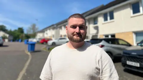 A man in a white tshirt with short brown hair and a beard stands in a residential street