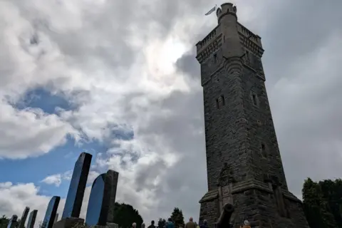 A view up towards the top of the MacDonald Monument. It is a tall, stone tower with narrow windows, and faux turrent and battlements. It is a sunny day.
