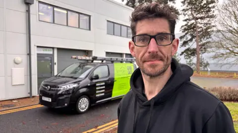 Nigel Day, wearing a black hoodie, in front of one of his heat pump installation firm's black and green vans 