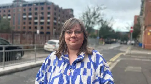 Terri McCurley smiling at the camera with shoulder length brown hair and a side fringe. She is wearing a blue and white print zip-up fleece and glasses.