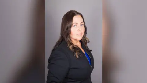 Pictured is Joanne Monk standing to the side with her head facing the camera. She has shoulder length brown hair
