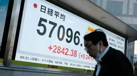 AFP via Getty Images A man walks in front of a giant screen showing the Nikkei Index at 57096