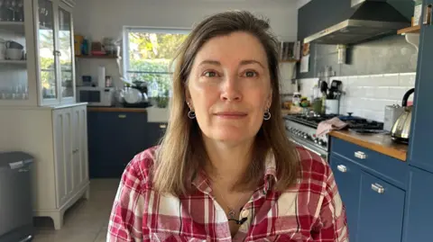 A woman sitting in her kitchen wearing a red checked shirt with blue kitchen cupboards behind her