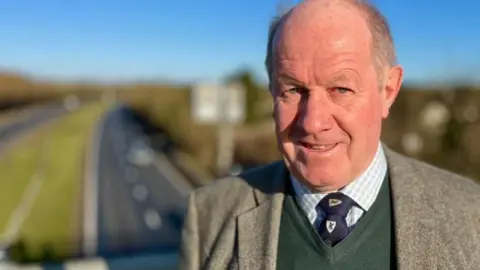Martin Giles/BBC Tim Passmore with very short grey hair, smiling at camera, wearing a grey jacket, green sweater and white shirt with blue tie which has a white loco. He is standing on a road bridge - cars are visible on the road behind him.