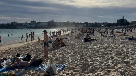 BBC/Tessa Wong A picture of Bondi Beach as the sun is starting setting. People can be seen sitting on towels and at the edge of the water. Buildings can be seen int he background