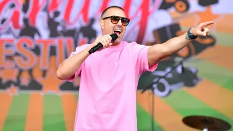 JCB Paddy McGuinness is on a stage wearing sunglasses and a pink shirt. He is holding a microphone up to his mouth, which is wide open, as he looks and points to his left. 