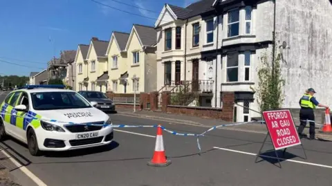 A police car is seen along a road, with police tape and a red "Road Closed" sign