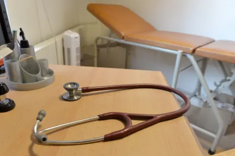 A brown stethoscope placed on a doctor's desk with a medical bed in the background