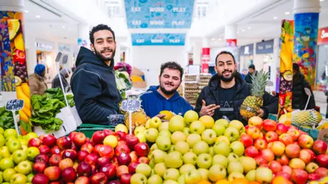 Fruity Bliss Three men smiling widely behind apples at a fruit stand.